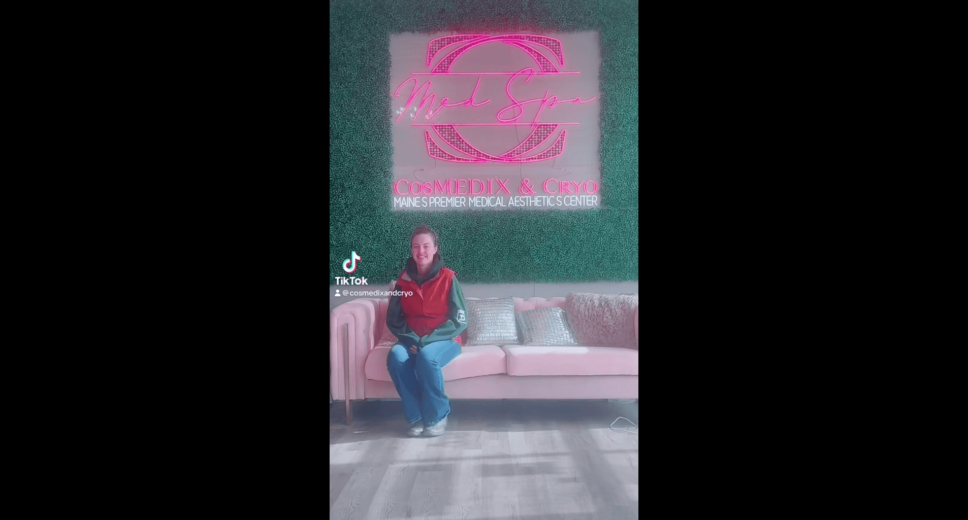 A woman sitting on the couch in front of a pink wall.